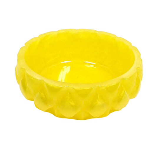 Angled side view of the handmade yellow resin ring dish. It is round with a carved like geometric design of raised diamond shapes around the outside. The resin is shiny and opaque in a bright pearly yellow color.