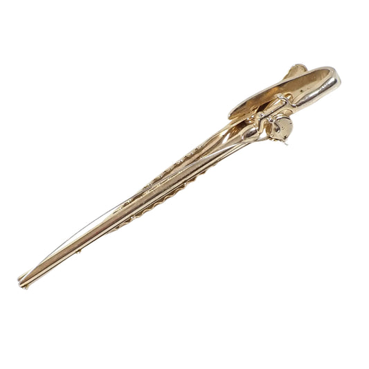 Angled view of the Hickok USA Mid Century vintage tie clip. The metal is gold tone in color. It is designed to look like a rod and open reel fishing pole and has a wire to depict the fishing line. It has an alligator clip on the back.
