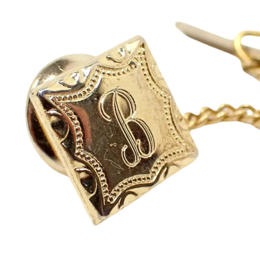 Enlarged view of the vintage initial tie tack. The metal is gold tone in color. There is an engraved fancy script letter B in the middle with a fancy dotted design around the edge of the tie tack.