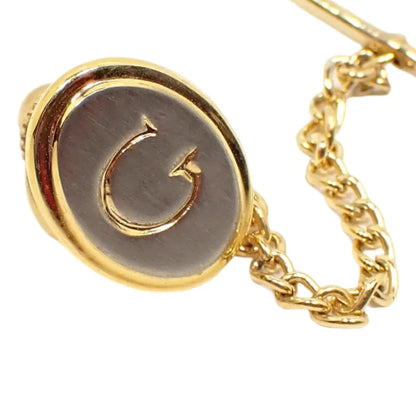 Enlarged view of the vintage initial tie tack. The front is silver tone in color with an engraved letter G in the middle in gold tone color. The rest of the tie tack, clutch, and chain are all gold tone in color.