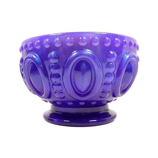 Side view of the handmade iridescent purple resin footed bowl. It is round and tapered at the bottom with a flared out footed area. The bowl part has large raised ovals going around it with a line of raised dots on top and bottom and in between each oval. The footed base is smooth. The resin is pearly iridescent with shades of purple and highlights of blue where the light is hitting it.