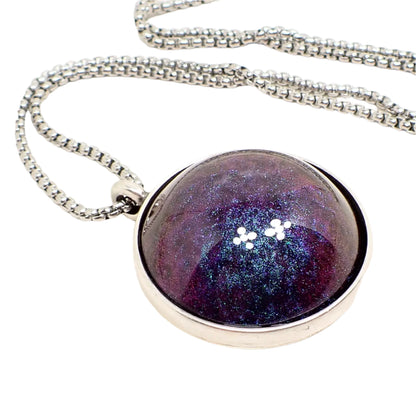 Enlarged close up of the pendant. It is round with an antiqued silver tone setting. The domed round resin cab has color shift resin that goes from dark purple to blue depending on how the light hits it.