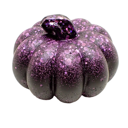 Angled view of the small pumpkin. It has matte black resin with iridescent purple glitter on top and in the stem of the pumpkin. The stem is the only shiny part of the piece, the rest has the matte appearance.