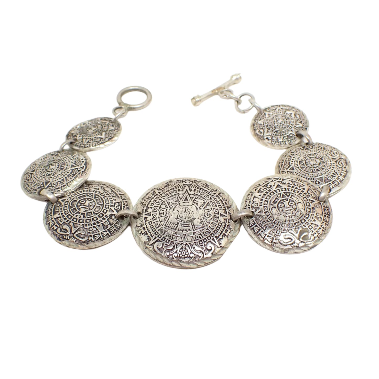 Angled top view of the vintage Mayan Calendar link bracelet. It has smaller to large round stamped disc links with depiction of Mayan Calendar on them. The largest link is in the middle of the bracelet. There is a toggle clasp at the end. The metal is antiqued silver tone in color.