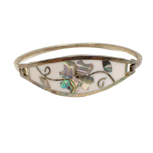 Front view of the Mexican Alpaca vintage hinged bangle bracelet. The metal has darkened from age and is a gray antiqued silver color. The front is white enameled and wider in the middle. It has an inlaid abalone shell flower design inthe middle with a leaf and stem on each side of it.