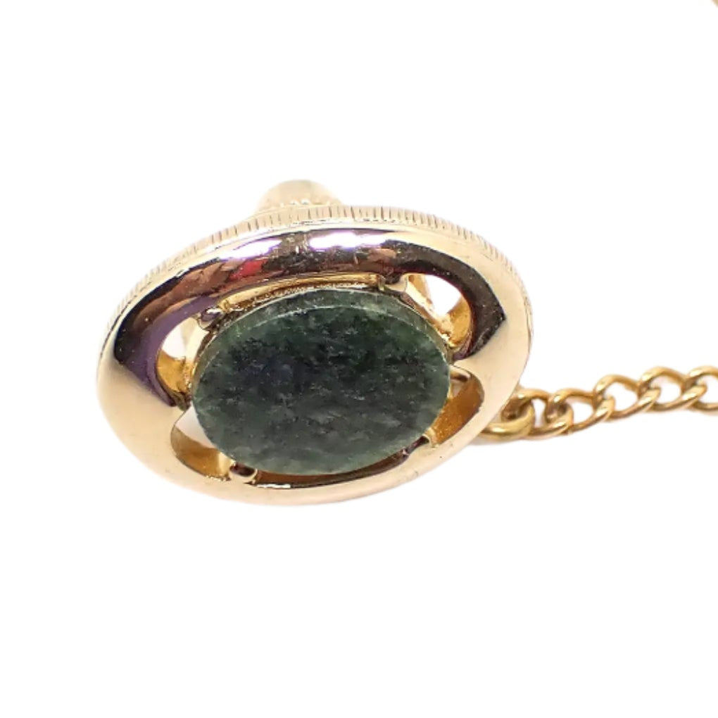 Enlarged view of the vintage tie tack. The metal is gold in color and has a Modernist oval design that has cut out areas and a green oval moss agate gemstone in the middle. Part of the clutch chain can be seen in the photo.