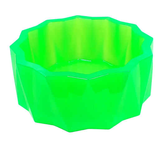 Angled side view of the large handmade neon green resin faceted bowl. The sides have a faceted triangle pattern that alternates in direction. The top of the bowl has a flat faceted sun shape design. The resin is shiny and semi translucent in a bright neon green color. Great for storing small items, as part of your table centerpiece, as a succulent planter pot, or just added to your UV fluorescent home decor.