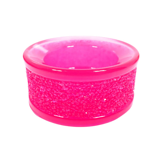 Angled side view of the handmade neon pink tealight candle holder. It is round with shiny semi translucent resin in a bright neon pink color. The top and bottom edge are rounded and smooth. The middle has a textured faux druzy design all the way around it that is sparkly when the light hits it. Fun piece to add to your home decor.