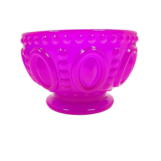 Side view of the handmade neon purple resin footed decorative bowl. It has a round bowl shape with tapered bottom and flared out foot. The bowl has large raised ovals all the way around it with lines of raised dots on top and bottom and in between the ovals. The foot is smooth. The resin is shiny and semi translucent in a bright neon purple color. Good for using as a small succulent planter pot, a small trinket dish, or just to add to your fluorescent home decor.