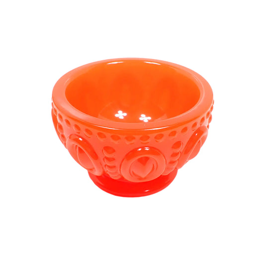 Angled side view of the handmade neon reddish orange resin decorative footed bowl. It is round with a tapered bottom with a flared round footed area. There are large raised ovals around the bowl with lines of raised dots above, below, and between the ovals. The resin is shiny and mostly opaque in a neon reddish orange color.