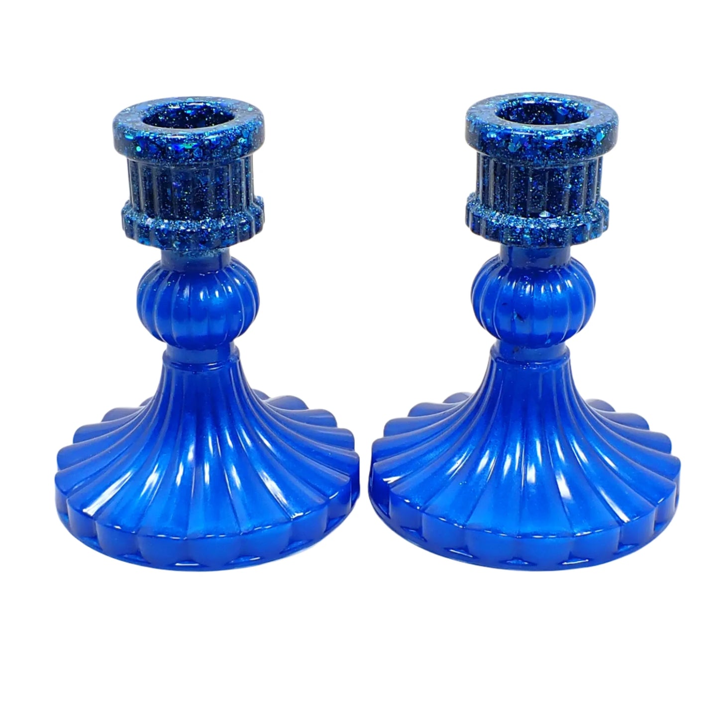 Angled side view of the ocean blue candle holders. There are two candlestick holders in a vintage style with corrugated round bottoms and tops. The tops have iridescent blue glitter and the rest is pearly ocean blue color resin.