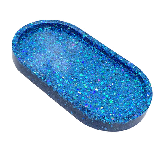 Angled view of the handmade resin ocean blue trinket dish. It is oval in shape and has chunky glitter with iridescent shades of ocean blue that sparkles in the light.