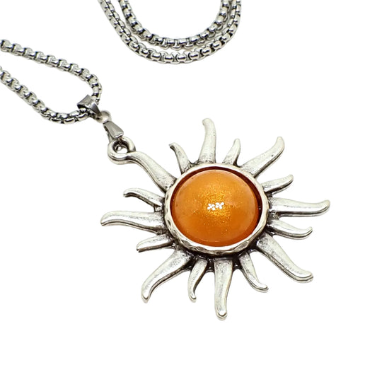 Enlarged close up photo of the sun pendant and part of the chain. The pendant is shaped like a sun with flames around it. The middle has a domed pearly orange resin cab.