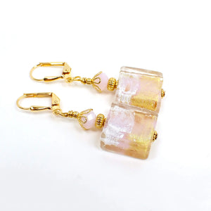 Angled view of the handmade drop earrings with fancy glass beads. The metal is gold plated in color. There are faceted opaque light pink glass crystals at the top. The bottom fancy glass beads are square shaped and have pink with areas of metallic gold, silver, and copper color.