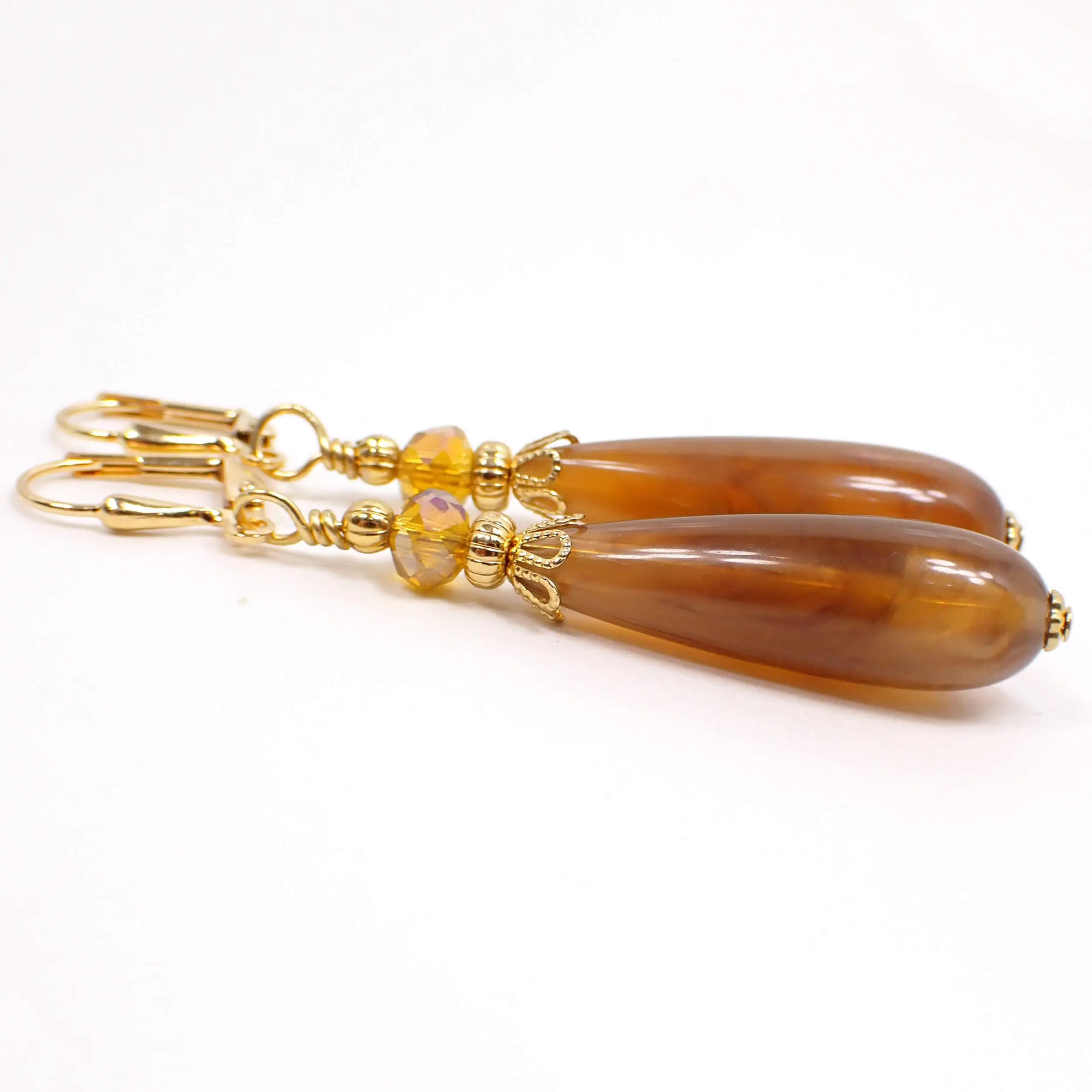 Side view of the handmade marbled lucite beaded earrings. The metal is gold plated in color. There is a faceted orange glass crystal bead at the top and a marbled vintage lucite teardrop bead at the bottom. The teardrop bead has swirls of caramel brown and orange colors.