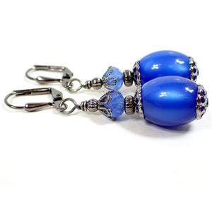 Side view of the handmade drop earrings with vintage beads. The metal is gunmetal gray in color. There are faceted glass beads in a semi translucent blue at the top. The bottom vintage beads are oval shaped moonglow lucite in a vivid blue color. They have an inner glow like appearance as you move around in the light.