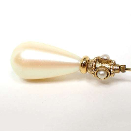 Enlarged top view of the retro vintage faux pearl stick pin. There is a golden off white color plastic teardrop shaped faux pearl at the top. Underneath that are small round faux pearls around the pin. The metal is gold tone plated in color.