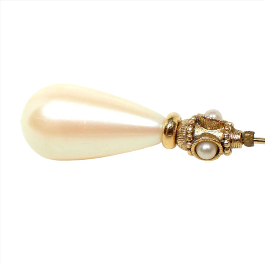 Enlarged top view of the retro vintage faux pearl stick pin. There is a golden off white color plastic teardrop shaped faux pearl at the top. Underneath that are small round faux pearls around the pin. The metal is gold tone plated in color.