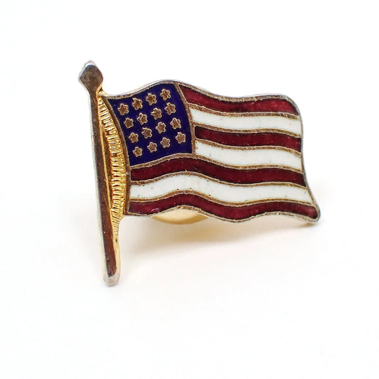 Front view of the retro vintage tie tack. It is shaped like a flag and has a US flag design with stars and stripes. The colors are enameled red, white, and blue. The metal is gold tone in color.