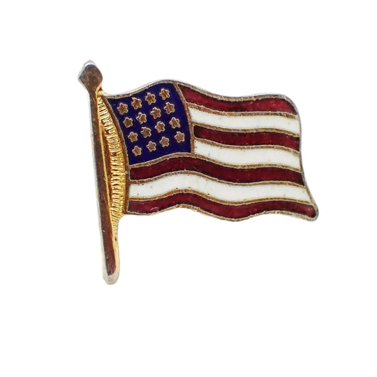Front view of the retro vintage tie tack. It is shaped like a flag and has a US flag design with stars and stripes. The colors are enameled red, white, and blue. The metal is gold tone in color.