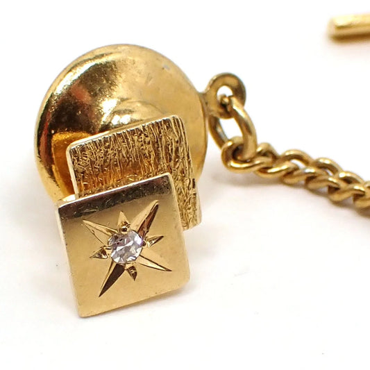 Angled view of the BAB Ballou Mid Century tie tack. The tie tack is 14K yellow gold and has a textured square in back and a shiny square in the front. The squares are angled so you see both. The gold plated clutch has a chain and bar on the end. The front of the shiny square has an etched atomic starburst design with a small diamond accent in the middle of it.