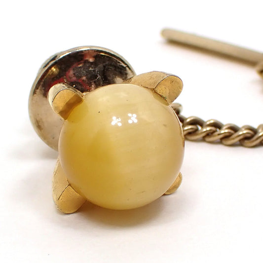Enlarged view of the Mid Century vintage faux cat's eye glass stud tie tack. The metal is gold tone in color. The round glass sphere in the middle is golden yellow in color and has a band of lighter color that seems to move as you move around in the light.