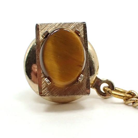 Front view of the Swank retro vintage gemstone tie tack. It is rectangle shaped with a brushed matte gold tone color setting. The gemstone on the front is an oval shaped tiger's eye cab with shades of yellow and brown. The back clutch and chain can be seen in the photo.