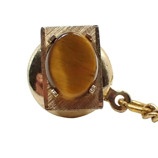 Front view of the Swank retro vintage gemstone tie tack. It is rectangle shaped with a brushed matte gold tone color setting. The gemstone on the front is an oval shaped tiger's eye cab with shades of yellow and brown. The back clutch and chain can be seen in the photo.