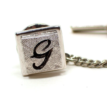 Enlarged closeup view of the Mid Century vintage Swank initial tie tack. It is square and silver tone in color. The front has a matte textured appearance with a cut out of the letter G in the middle. The metal behind the letter is painted black and shows through.