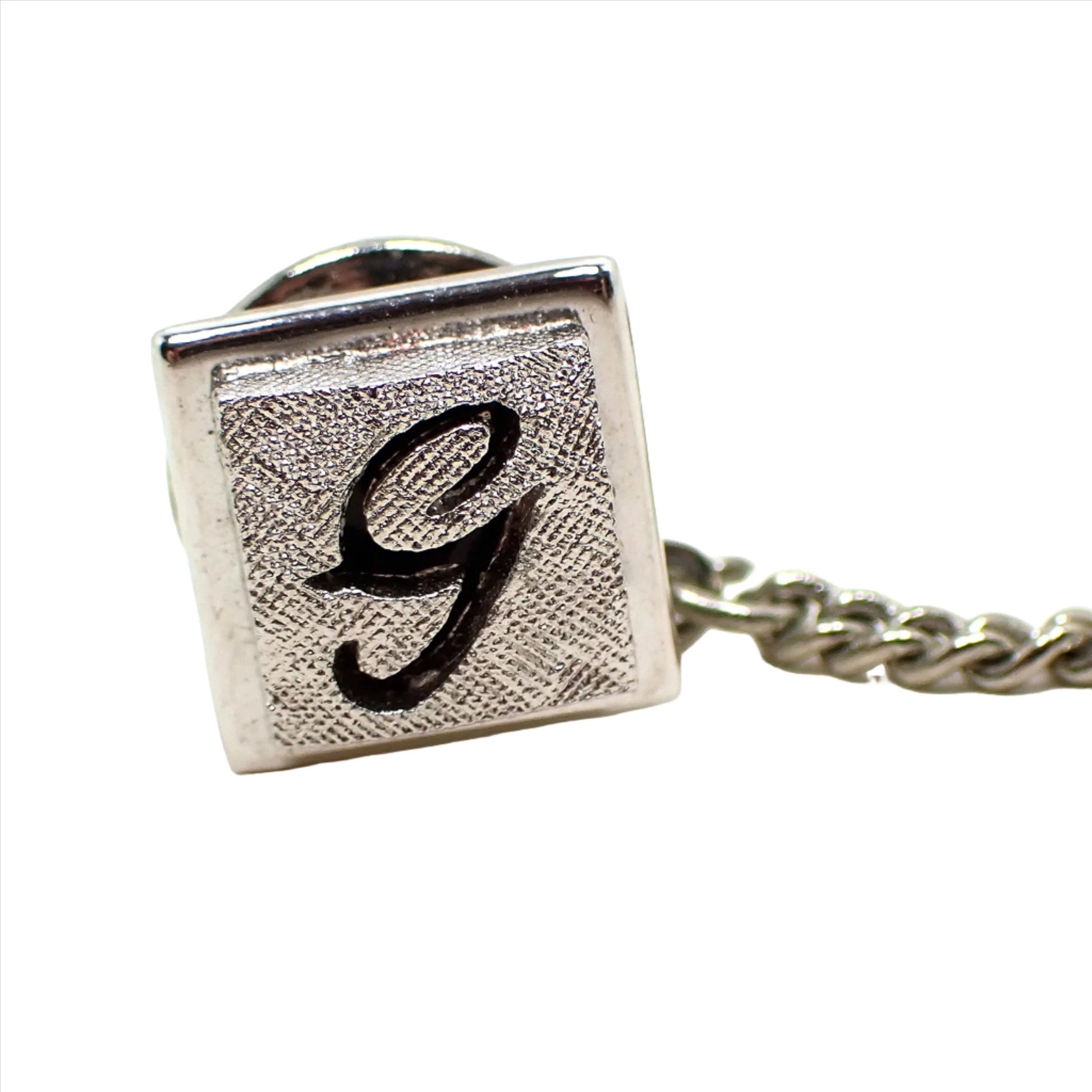 Enlarged closeup view of the Mid Century vintage Swank initial tie tack. It is square and silver tone in color. The front has a matte textured appearance with a cut out of the letter G in the middle. The metal behind the letter is painted black and shows through.