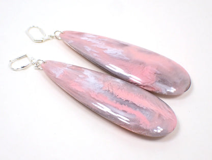 Angled view of the handmade large teardrop earrings. The metal is silver plated in color. There are very large long acrylic teardrops that have marbled pink and gray colors.