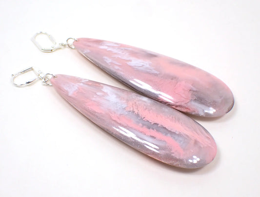 Angled view of the handmade large teardrop earrings. The metal is silver plated in color. There are very large long acrylic teardrops that have marbled pink and gray colors.