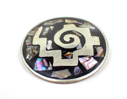 Enlarged front view of the retro vintage Mexican Southwestern brooch. It is round and has a silver tone tribal style design. The background has black enamel and there are various pieces of inlaid abalone shell that flash with different colors as you move around in the light. There are some small spot discolorations on the metal on the front when viewed under magnification.