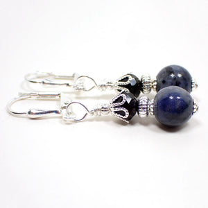 Side view of the small handmade sodalite gemstone earrings. The metal is silver plated in color. There are black faceted glass crystal beads at the top. The bottom gemstone beads are round sphere shaped and mostly dark denim blue in color with specks of black and gray.