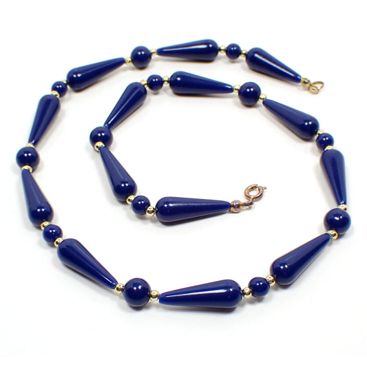 Top camera view of the retro vintage lucite beaded necklace. The meal is gold plated in color. There is a spring ring clasp at the end that is darkened from age. The necklace is beaded with dark blue lucite beads that alternate between teardrop and round shape with small round gold plated beads in between.