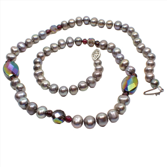 Top view of the retro vintage glass beaded faux pearl necklace. The imitation pearl beads are gray and purplish gray in color. In a few spots on the necklace there are small round dark purple beads with a faceted AB bead in between. There is a filigree push lock clasp on the end in silver color that is white gold filled.