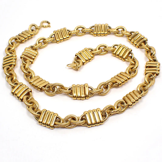 Top view of the retro vintage Trifari fancy link chain necklace. The metal is gold tone plated in color. There is a spring ring clasp at the end. The necklace has a repeating pattern of three textured oval cable links and then a link that is long with four oval bar link links over open links that stick out on each end. 