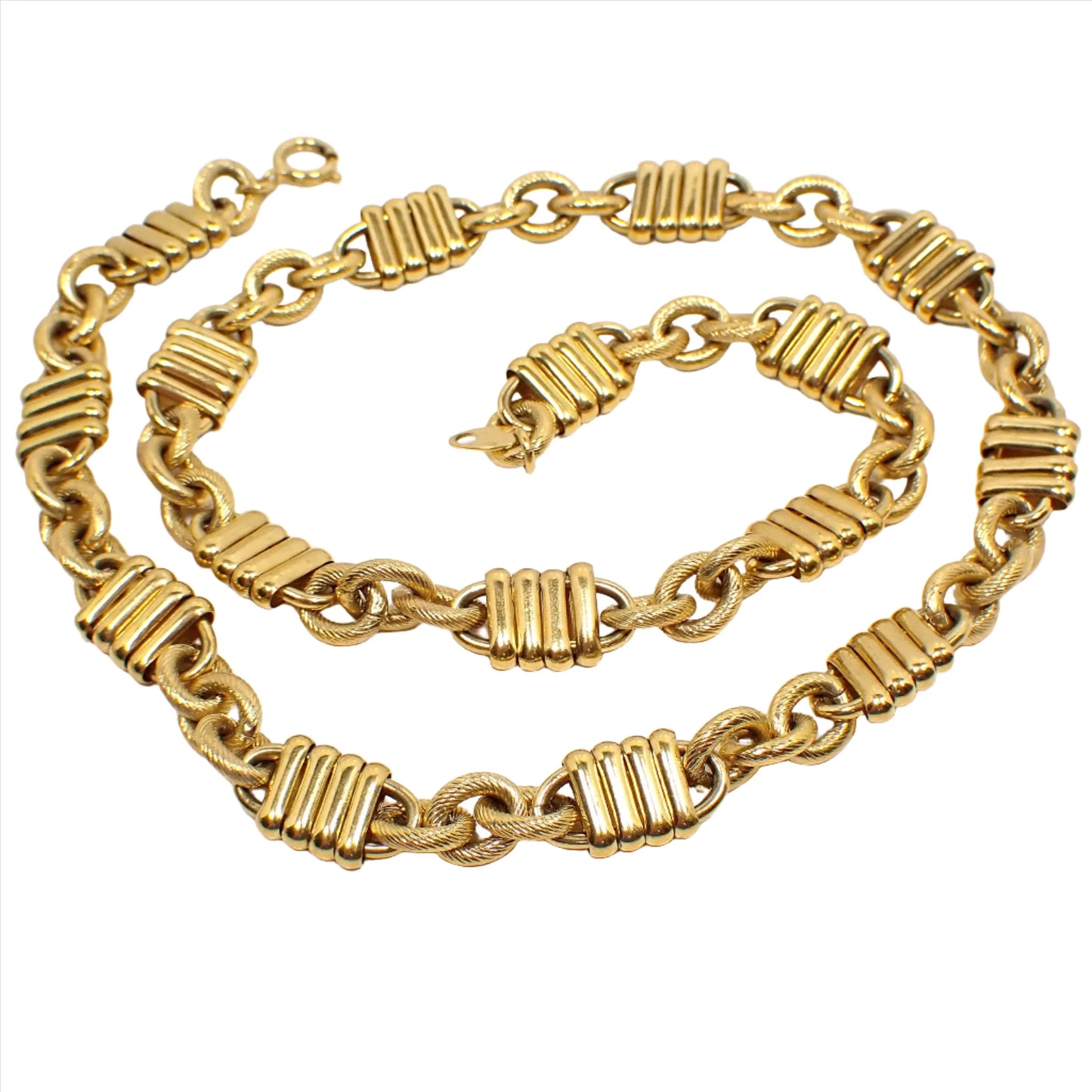 Top view of the retro vintage Trifari fancy link chain necklace. The metal is gold tone plated in color. There is a spring ring clasp at the end. The necklace has a repeating pattern of three textured oval cable links and then a link that is long with four oval bar link links over open links that stick out on each end. 