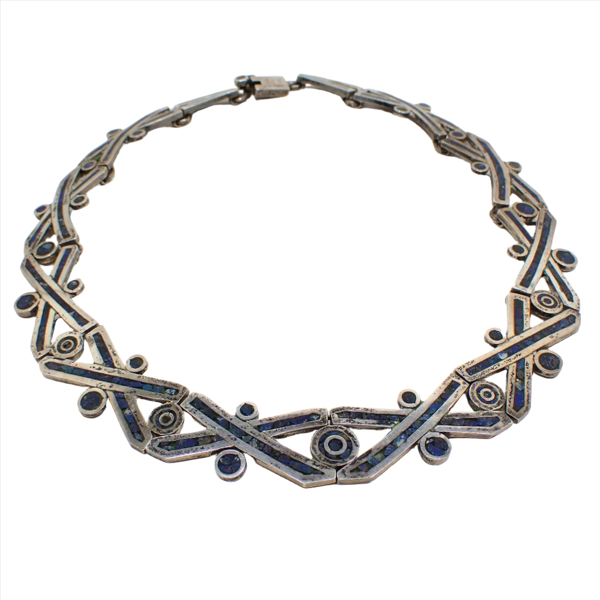 Angled front view of the retro vintage Taxco sterling silver link necklace. The necklace has longer style links that are shaped like X's with a dot above, below, and in between. Each piece has inlaid lapis lazuli gemstone chips that are dark blue in color. The sterling silver is darkened from age for a gray color with some small areas of black. There is a slide lock clasp at the end.