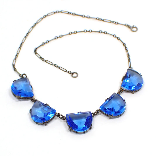 Top view of the 1930's Art Deco Czech glass necklace. The sterling silver chain and settings are darkened from age to a gray color. There are five scalloped semi circle drops that have faceted blue glass cabs. Each one is in a bezel setting with prongs to hold them in and they have an open back design. There is a spring ring clasp at the end.