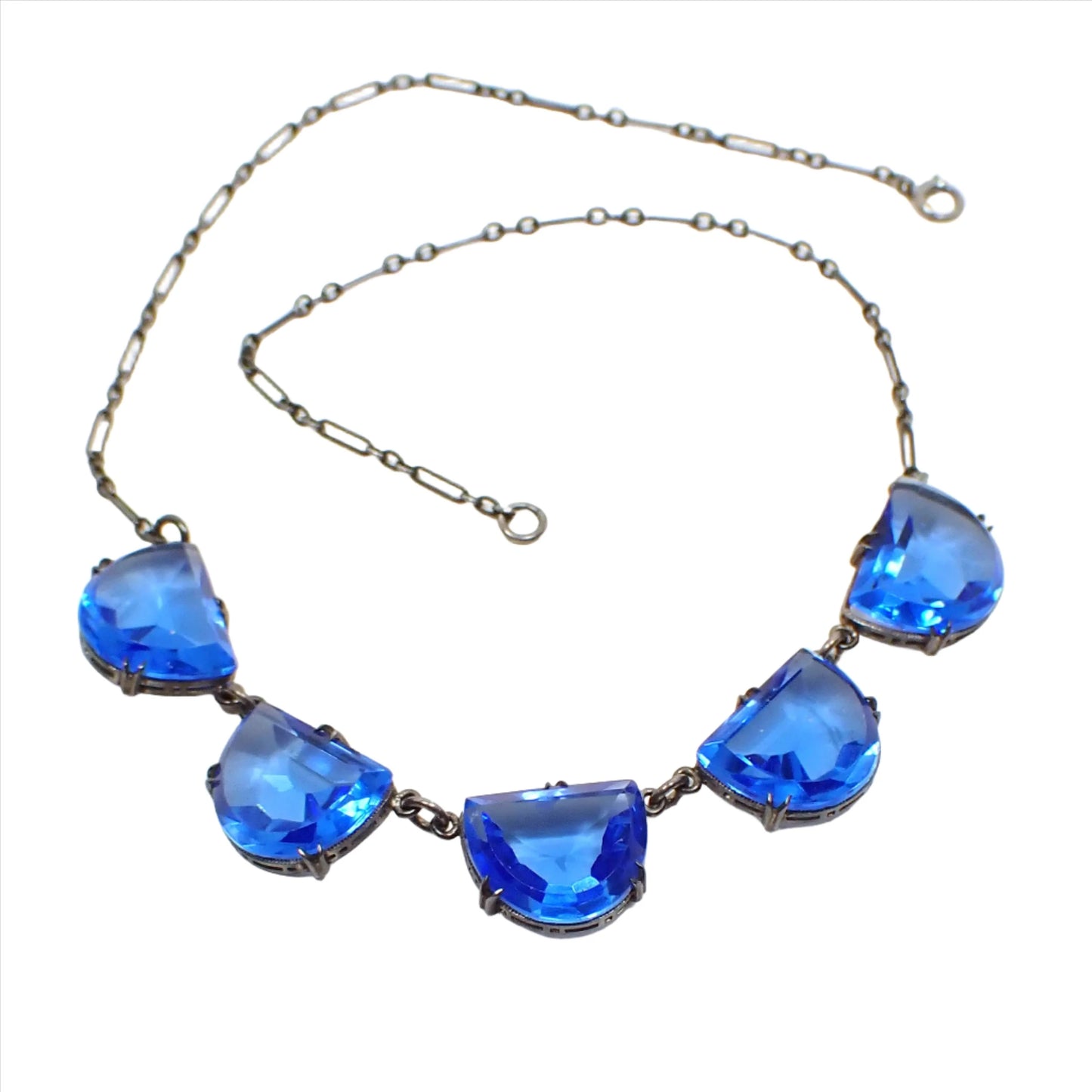 Top view of the 1930's Art Deco Czech glass necklace. The sterling silver chain and settings are darkened from age to a gray color. There are five scalloped semi circle drops that have faceted blue glass cabs. Each one is in a bezel setting with prongs to hold them in and they have an open back design. There is a spring ring clasp at the end.