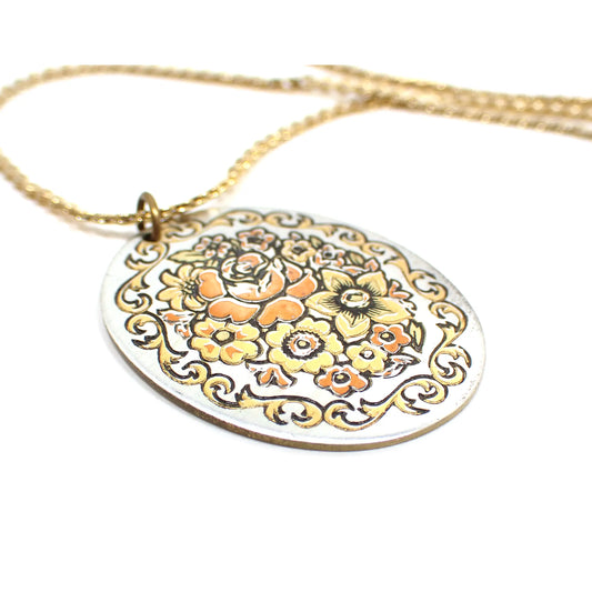 Enlarged view of the retro vintage Reed and Barton Damascene pendant necklace. The chain is gold tone in color. The pendant is flat oval shaped and is silver tone in color. There are various flowers etched on the front in gold and copper colors and a gold tone curvy leaf like border around the edge.