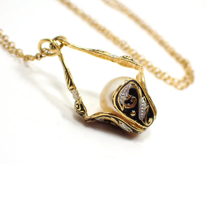 Enlarged view of the retro vintage Damascene style basket pendant necklace with faux pearl. The chain is a thinner style cable link. The pendant is shaped like a flower with rounded edges. The outside of the pendant has a Damascene style design with curly leaf type shapes and black and silver paint on the gold plated setting.