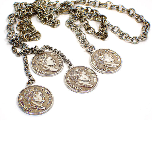 Enlarged view of the retro vintage faux coin medallion lariat necklace. The metal is mostly silver tone plated in color, but there is some darkened patina on the chain and faux coins from age. The chain is cable link with double rings and each link has one ring that is textured. This photo is showing the front view of the medallions with a depiction of Napoleon and the words Napoleon Empereur around the sides.