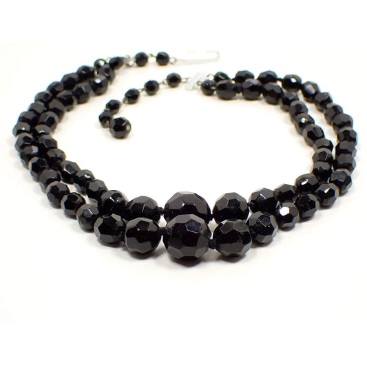 Photo of the Mid Century vintage beaded multi strand necklace from Germany. There are two strands of black faceted glass beads that are graduated in size with the largest being at the bottom of the necklace. There is a silver color aluminum hook clasp and beaded extender chain at the end.
