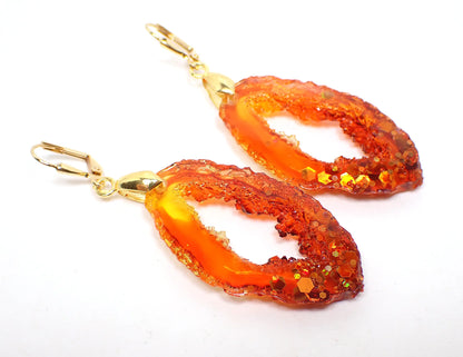 Angled front view of the handmade faux druzy geode slice style earrings. The metal is gold plated in color. The resin is mostly fiery red and orange with a hint of bright yellow at the top. There is chunky iridescent glitter at the bottom with shades of orange and yellow.
