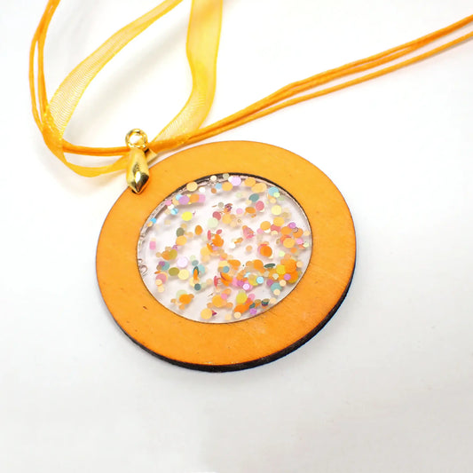 Enlarged front view of the handmade wood and resin pendant. Part of the necklace is showing and has three strands of faux leather round cord and a flat organza ribbon strand. They are orange in color. The pendant is a flat round piece of orange dyed wood with an open middle area that is filled with multi color pastel confetti resin. The pendant bail is gold plated in color.