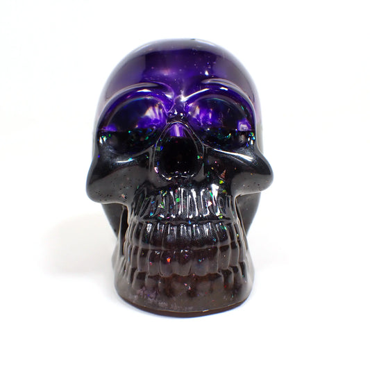 Front view of the handmade resin skull. The top part is dark semi translucent purple. The bottom has dark pearly gray and purple resin with areas of iridescent chunky glitter here and there.