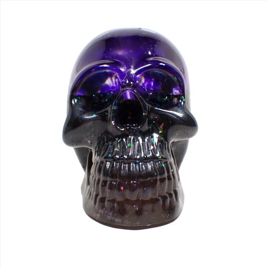 Front view of the handmade resin skull. The top part is dark semi translucent purple. The bottom has dark pearly gray and purple resin with areas of iridescent chunky glitter here and there.