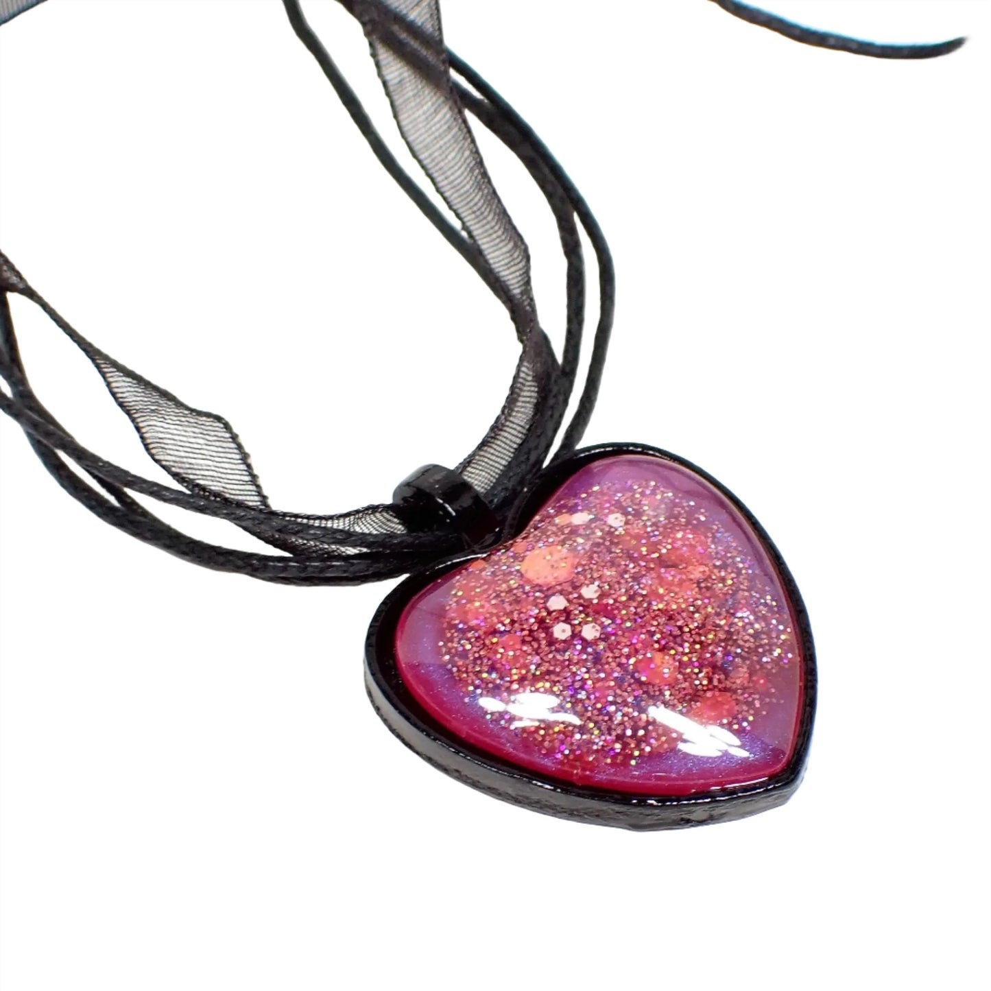 Enlarged view of the handmade black heart Goth pendant necklace. The necklace has three strands of faux leather cord and a strand of organza ribbon. The heart has black coated metal with a resin cab. The resin is bright pearly pink with hints of purple and chunky iridescent pink glitter.